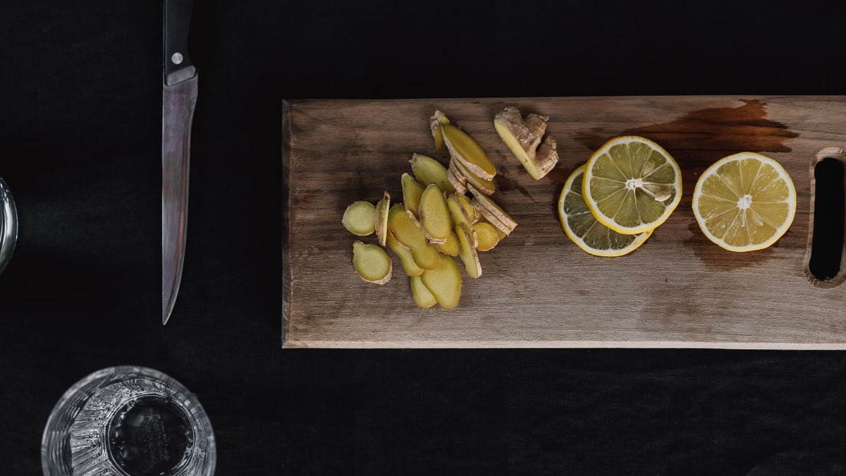 Top-down view of sliced ginger and lemons on a wooden chopping board with knife and glass.
